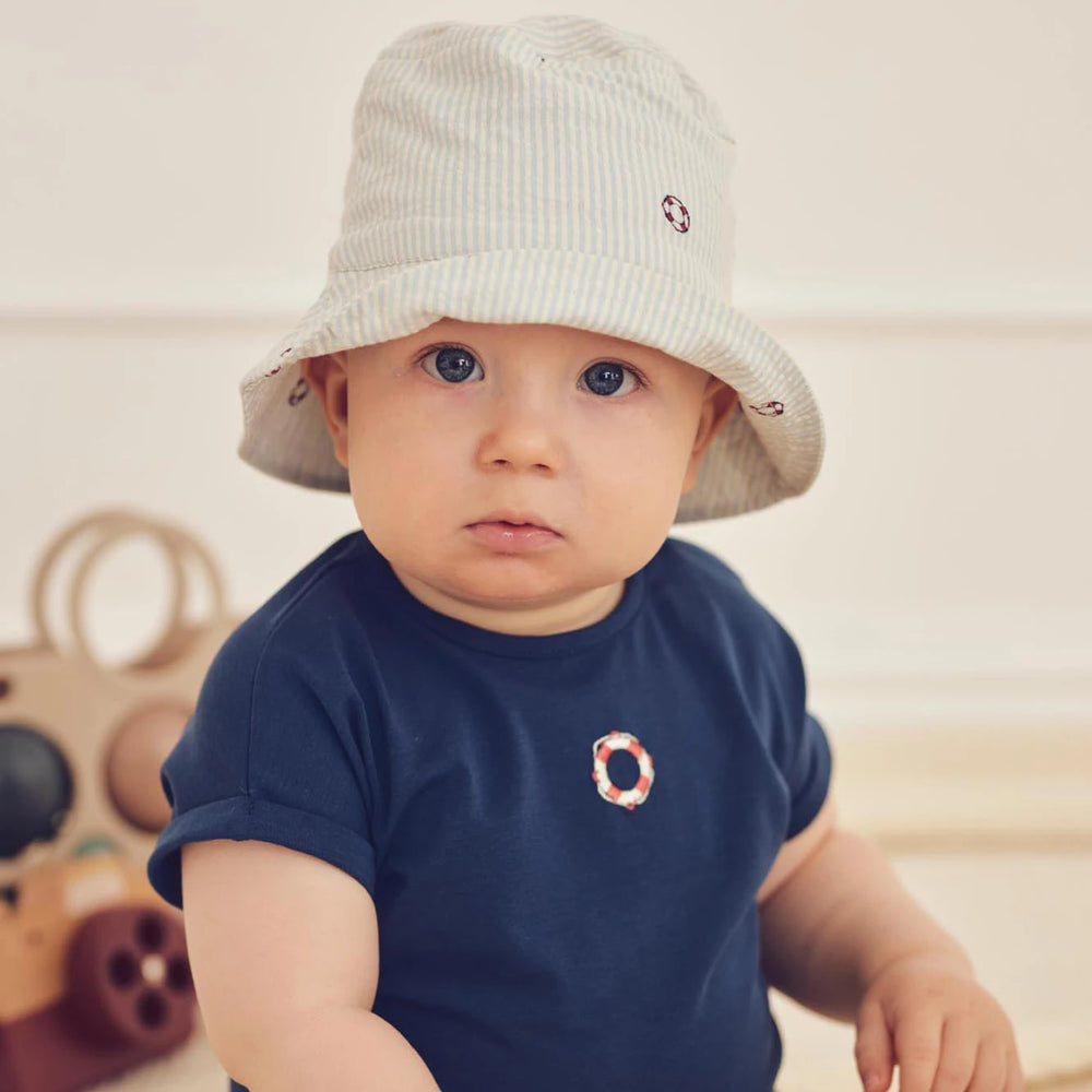 Baby wearing a white bucket hat and navy shirt with a circular design, sitting on a wooden floor.