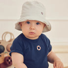 Baby wearing a white bucket hat and navy shirt with a circular design, sitting on a wooden floor.