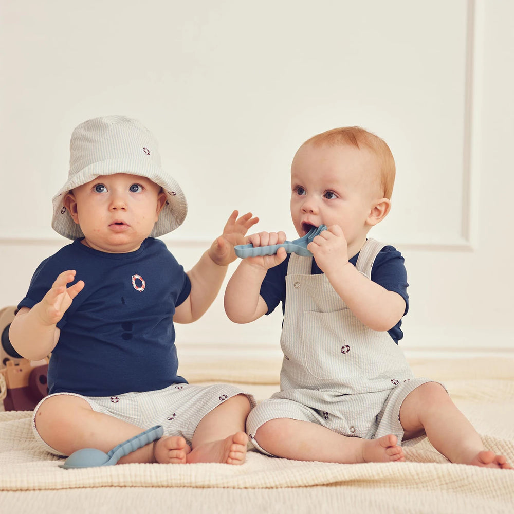 Two babies sitting on a blanket, one wearing a hat and the other holding a toy.