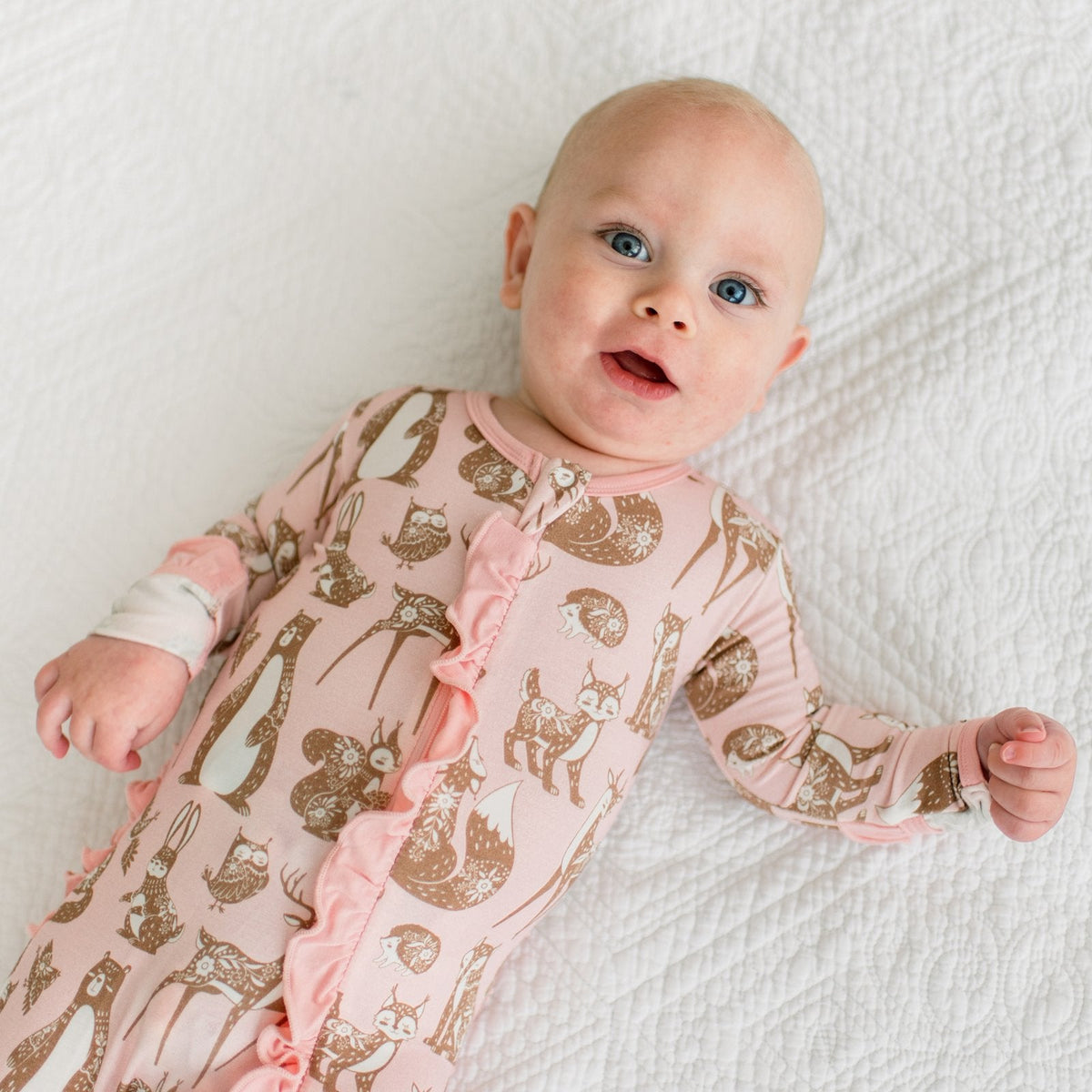 A baby lying on a white quilted blanket, wearing a light pink long-sleeve footless sleeper with a brown woodland animal print and ruffle trim down the front.
