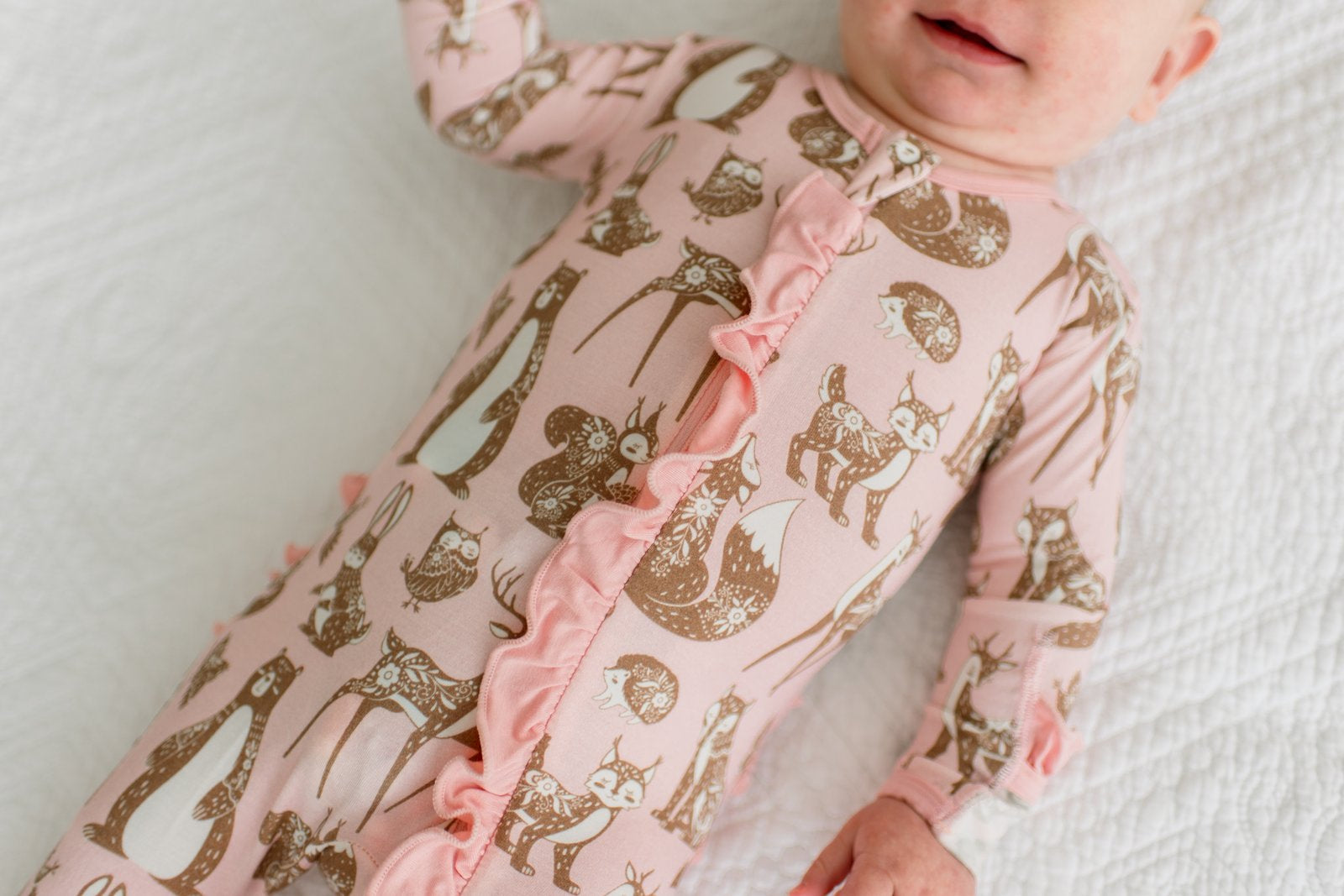 A baby lying on a white textured blanket, wearing a pink long-sleeve footless sleeper with a brown woodland animal print and ruffle trim down the front.