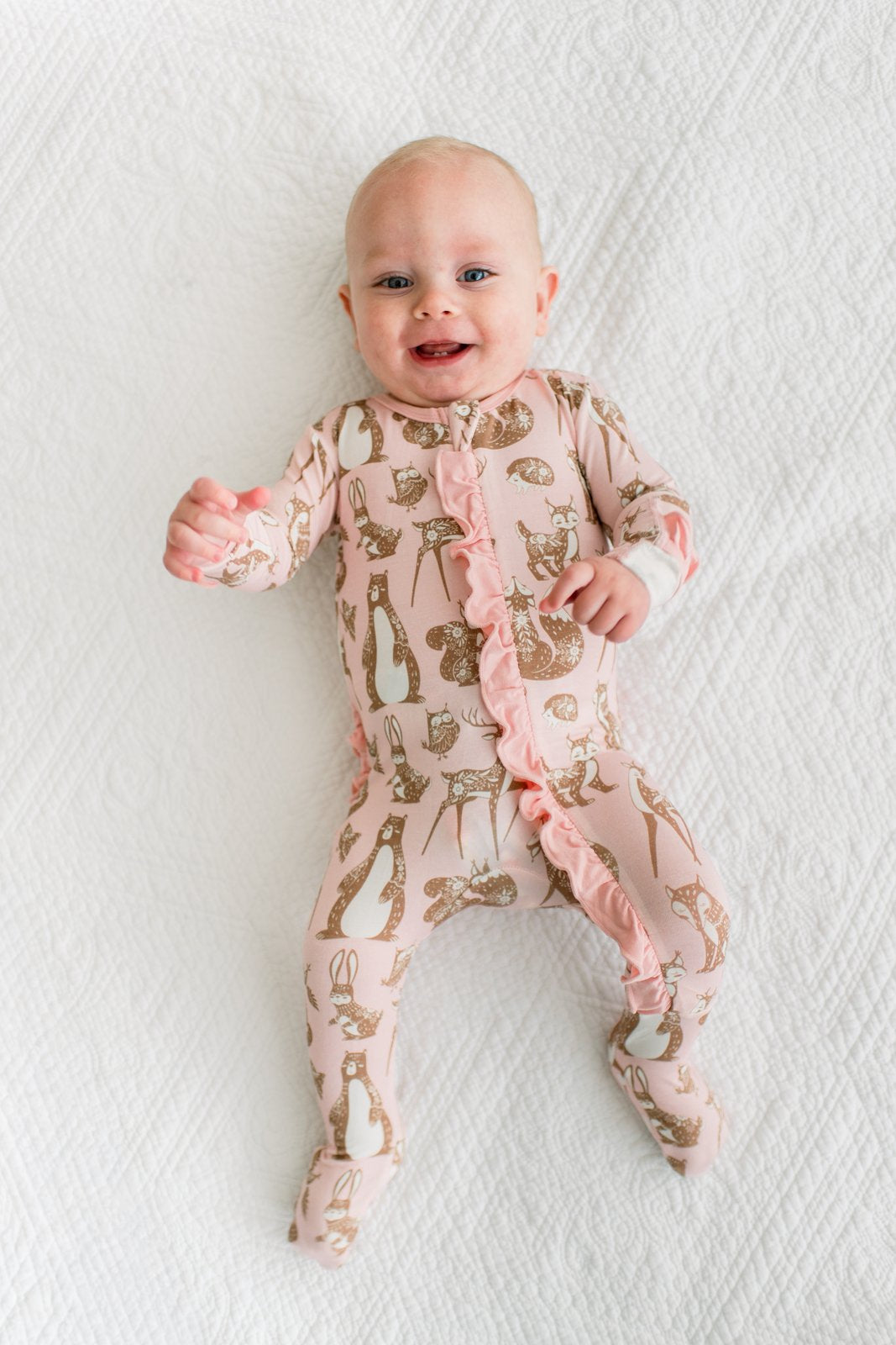 A baby lying on a white textured blanket, wearing a light pink footed sleeper with a gold animal print and ruffled trim.