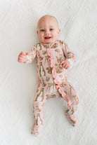 A baby lying on a white textured blanket, wearing a light pink footed sleeper with a gold animal print and ruffled trim.