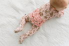 A baby lying on their stomach on a white textured blanket, wearing pink footed pajamas with a brown animal print and a ruffled waistband.