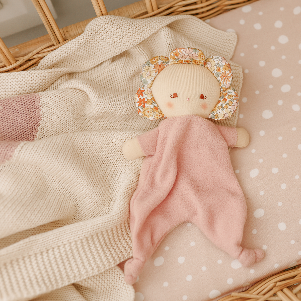 A soft baby lovey security blanket featuring a pale pink footed body and a floral-patterned bonnet, resting on a cream knit blanket inside a wicker basket.