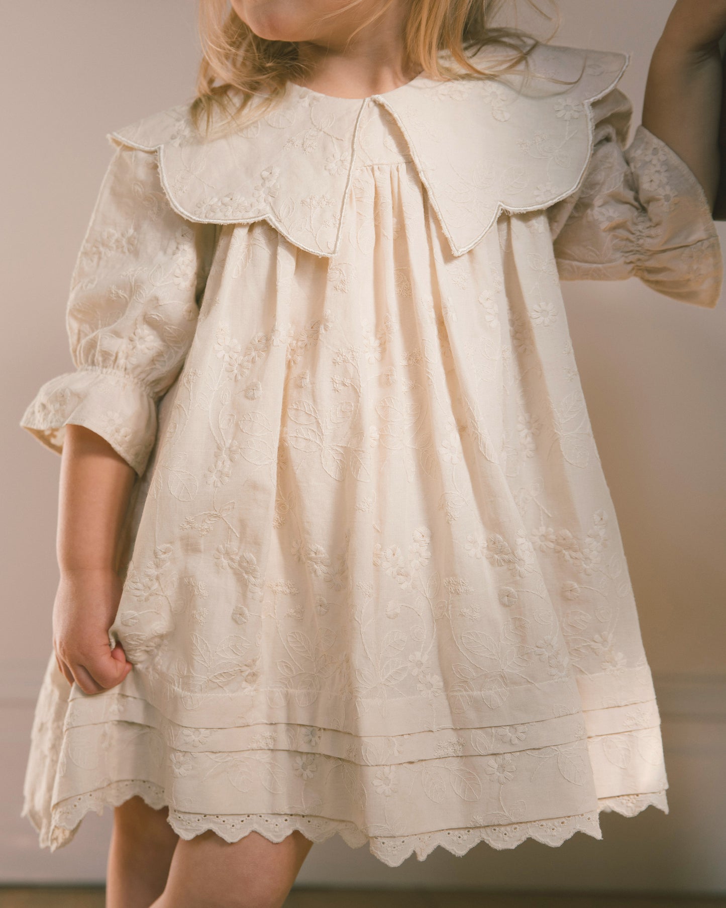 A cream-colored baby or toddler dress with a large ruffled Peter Pan collar, long puff sleeves, and delicate floral embroidery, shown on a child against a neutral background.