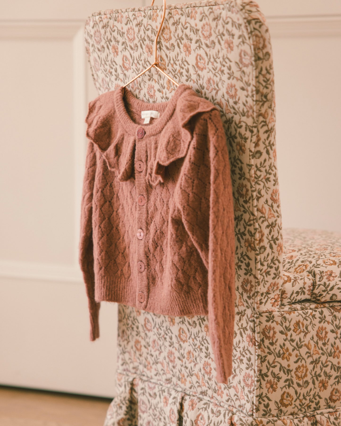 A dusty rose knit cardigan sweater with a ruffled collar and button front, hanging on a hanger from a floral-patterned chair in a softly lit indoor setting.
