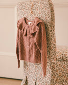 A dusty rose knit cardigan sweater with a ruffled collar and button front, hanging on a hanger from a floral-patterned chair in a softly lit indoor setting.