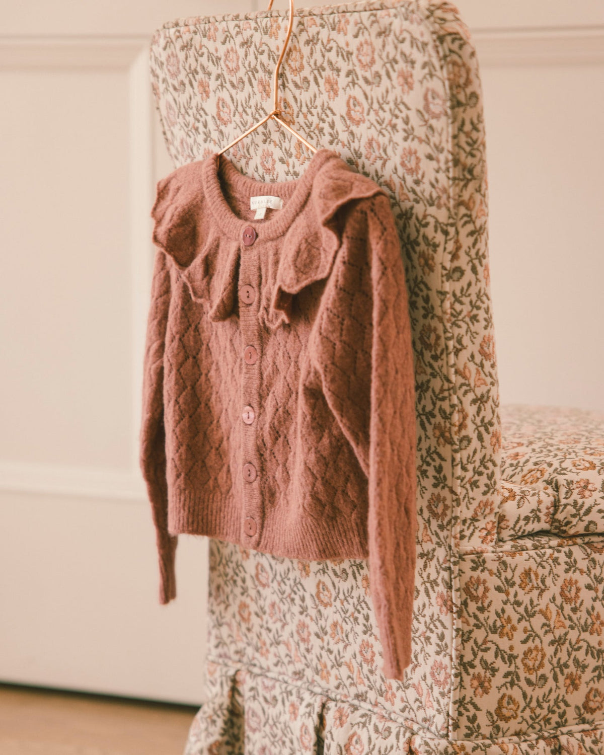 A dusty rose knit cardigan sweater with a ruffled collar and button front, hanging on a hanger from a floral-patterned chair in a softly lit indoor setting.