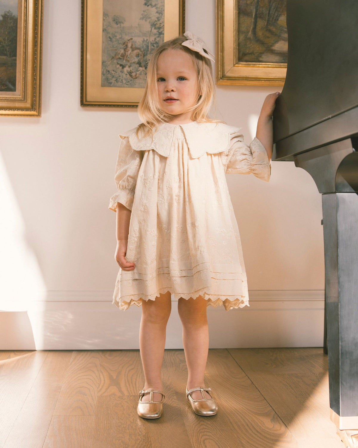 A young girl with blonde hair wearing a cream-colored lace dress and metallic gold shoes stands in a room next to a black grand piano, looking off-camera.
