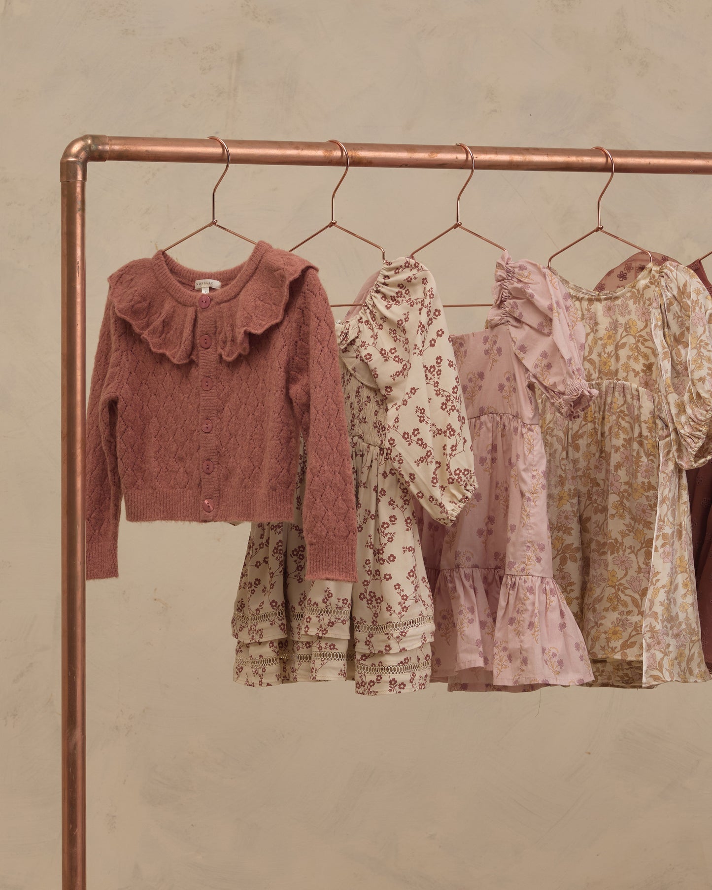 A copper-colored clothing rack displaying a collection of children's clothing, including a dusty rose knit cardigan with a ruffled collar and three floral dresses in cream, pink, and yellow tones, set against a textured beige wall.
