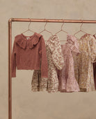 A copper-colored clothing rack displaying a collection of children's clothing, including a dusty rose knit cardigan with a ruffled collar and three floral dresses in cream, pink, and yellow tones, set against a textured beige wall.