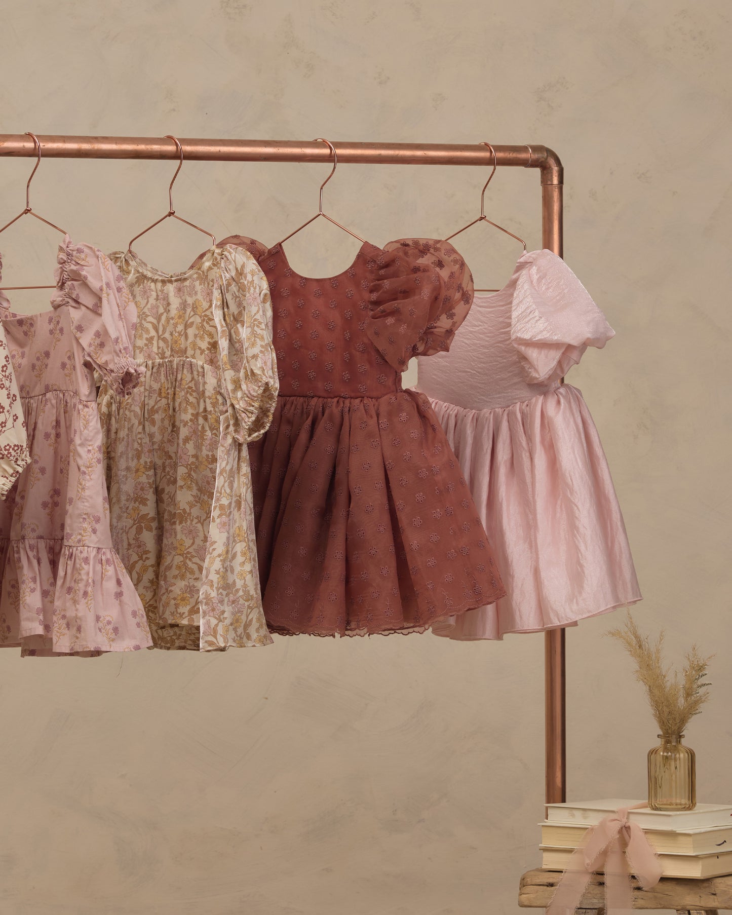 Four girls' dresses in soft pastel and muted tones hang on a copper clothing rack against a textured beige wall. The dresses include floral prints and solid fabrics in pink, cream, rust, and light pink.