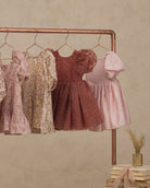 Four girls' dresses in soft pastel and muted tones hang on a copper clothing rack against a textured beige wall. The dresses include floral prints and solid fabrics in pink, cream, rust, and light pink.