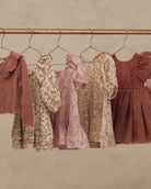 Four children's dresses in soft dusty pink, beige floral print, and rust tones hanging on a copper-colored clothing rack against a textured beige wall.