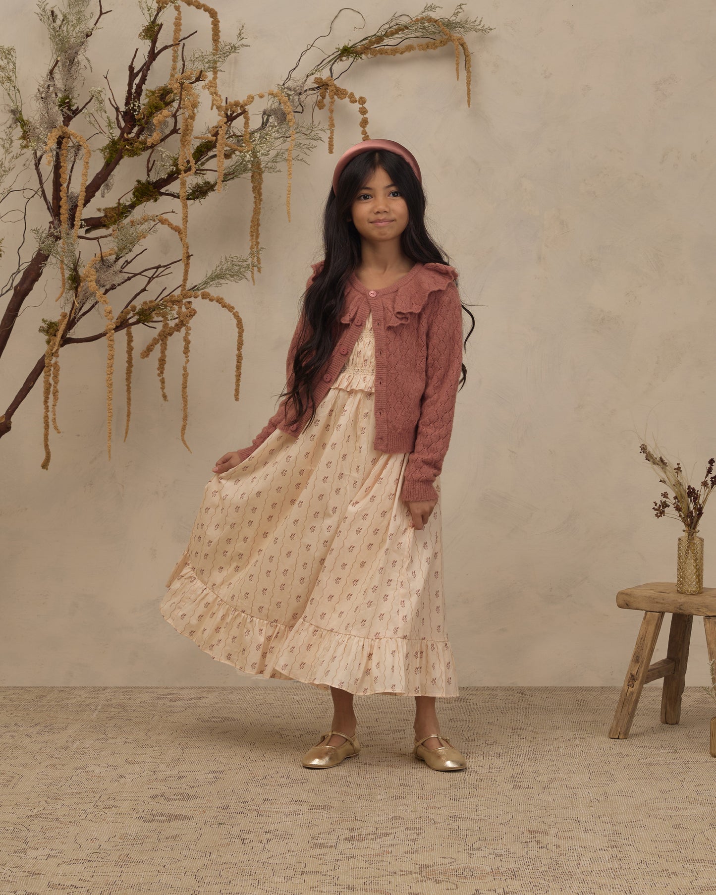 A young girl stands in a studio setting wearing a long cream dress with small pink floral prints, a dusty rose cardigan, and gold shoes. She is posed in front of a textured beige wall with dried botanical branches and a small wooden stool.