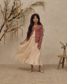 A young girl stands in a studio setting wearing a long cream dress with small pink floral prints, a dusty rose cardigan, and gold shoes. She is posed in front of a textured beige wall with dried botanical branches and a small wooden stool.