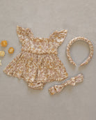 A baby girl's floral romper set in a beige and brown velvet-like fabric, styled flat-lay with a matching headband and a small bow headband on a textured beige background.