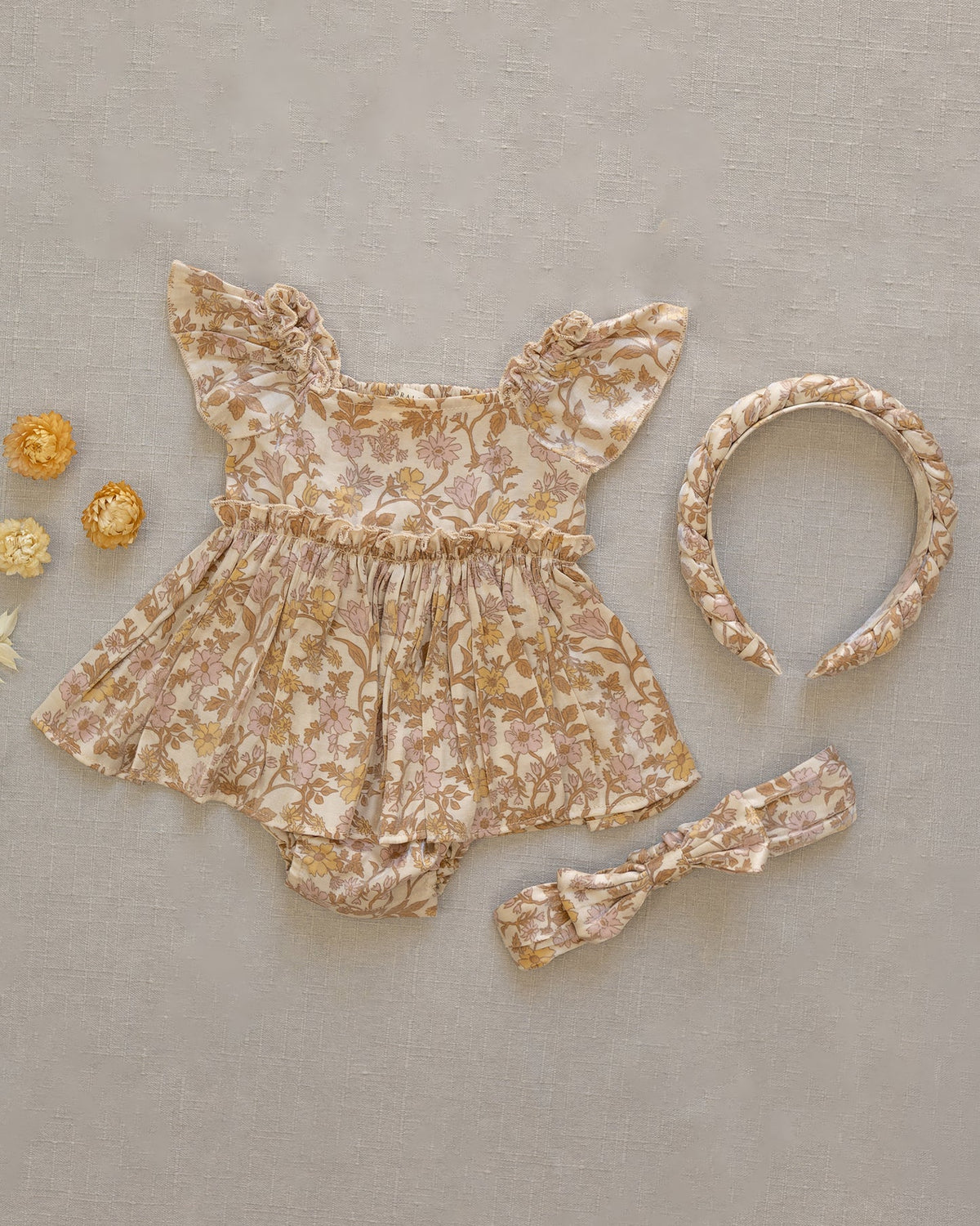 A baby girl's floral romper set in a beige and brown velvet-like fabric, styled flat-lay with a matching headband and a small bow headband on a textured beige background.