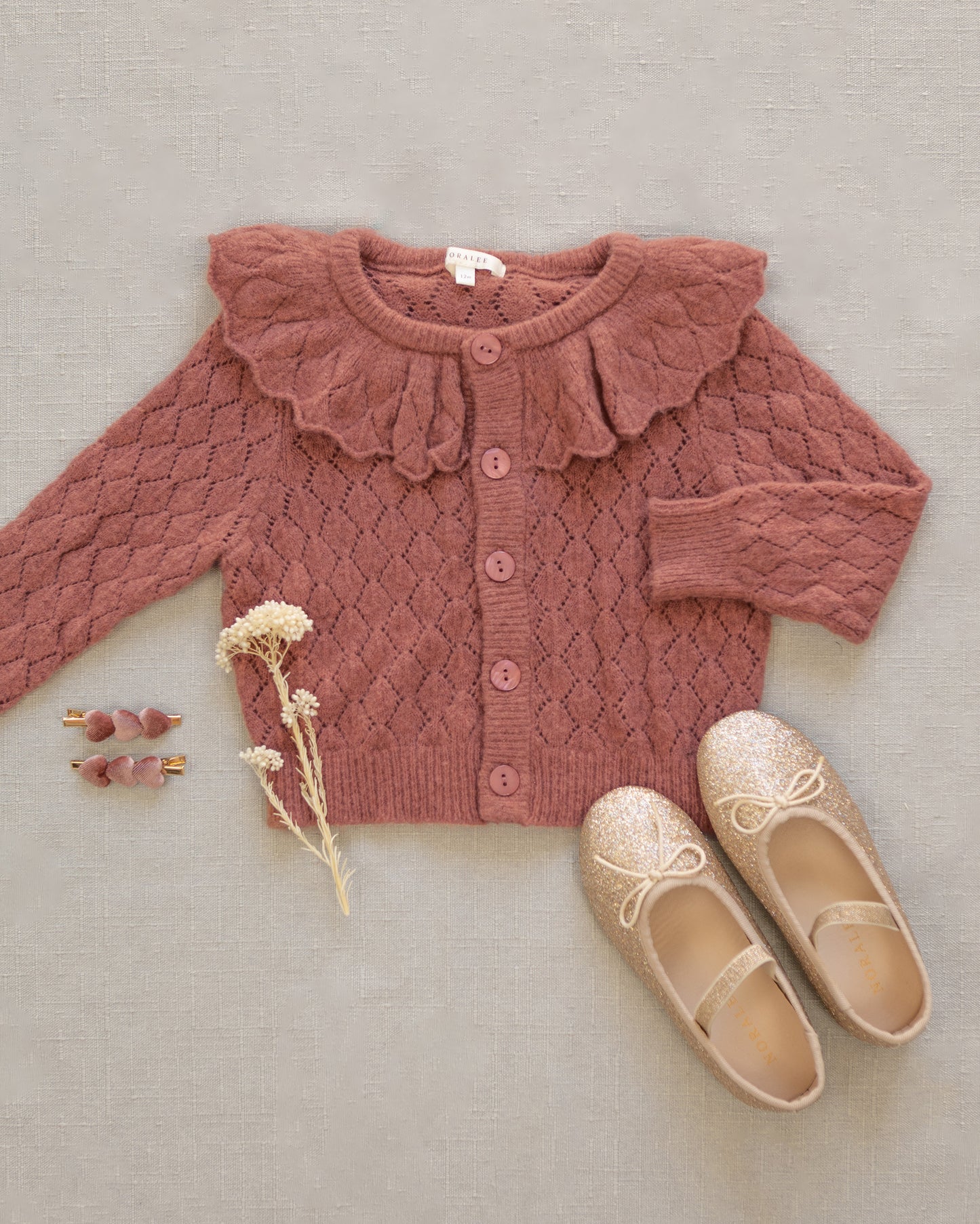 A dusty rose pink cable-knit baby cardigan with a large ruffled collar and button front, styled in a flat lay with beige glittery ballet flats, small heart-shaped earrings, and dried flowers on a textured beige fabric background.