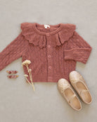 A dusty rose pink cable-knit baby cardigan with a large ruffled collar and button front, styled in a flat lay with beige glittery ballet flats, small heart-shaped earrings, and dried flowers on a textured beige fabric background.