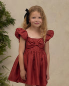 A young girl with blonde curly hair wearing a rust-red dress with puff sleeves, a bow at the neckline, and a black hair bow, standing against a textured beige wall.
