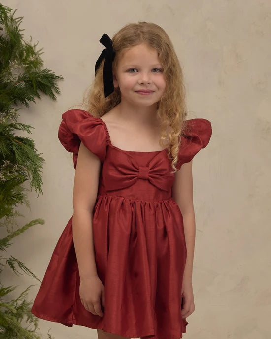 A young girl with blonde curly hair wearing a rust-red dress with puff sleeves, a bow at the neckline, and a black hair bow, standing against a textured beige wall.