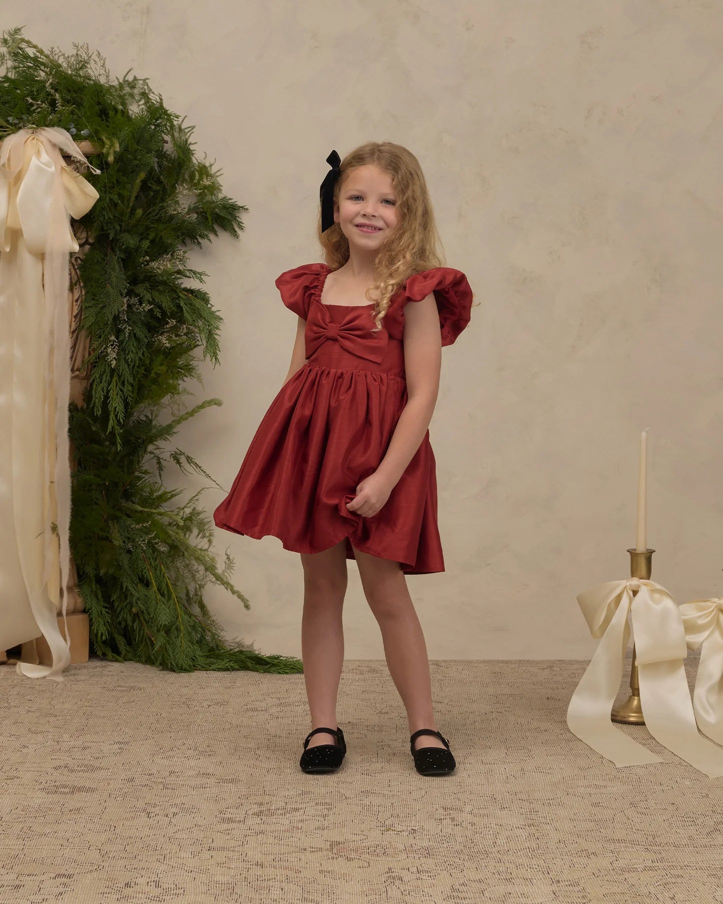 A young girl with blonde hair wearing a rust-red ruffled dress and black shoes, standing in a styled studio setting with a textured beige wall, greenery, and cream ribbons.