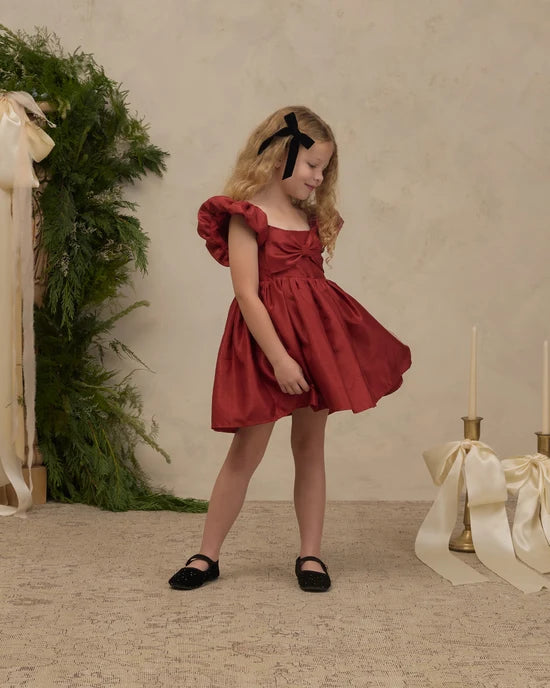 A young girl with blonde hair tied back with a black ribbon bow is wearing a short, red dress with ruffled sleeves and black Mary Jane shoes, standing in a styled studio setting with a textured beige wall, greenery, and decorative candles.