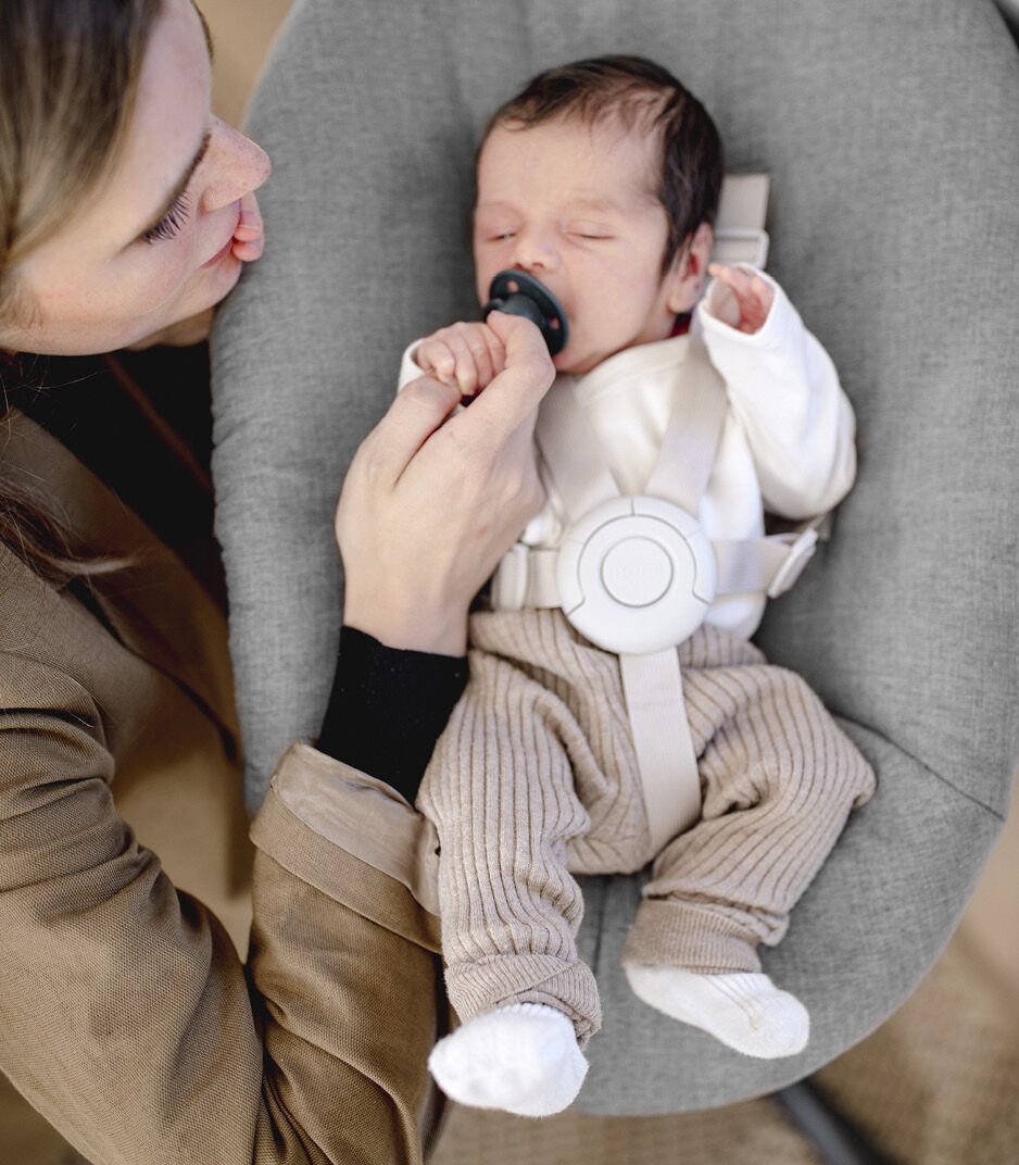 Newborn baby in a gray bouncer with a woman holding the baby's hand.
