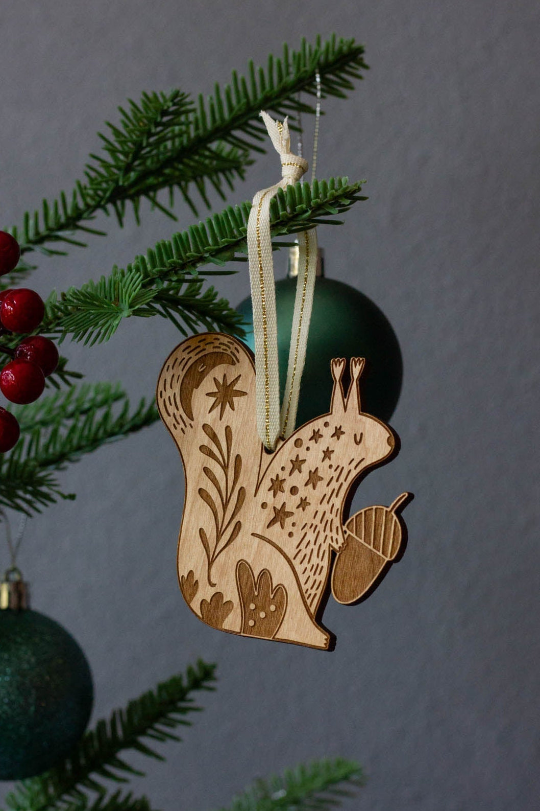 Wooden squirrel ornament hanging on a Christmas tree with green baubles and red berries.