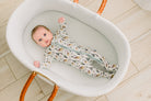 A baby lies on their back inside a white woven bassinet, wearing a long-sleeve footed sleeper with a white background and a pattern of brown and gray animals, featuring a light blue zipper.