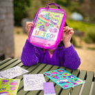 Person holding a 'Let's Go!' activity and coloring kit on a table with coloring sheets and stickers.