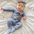 Baby in a blue outfit with a teething necklace lying on a white blanket.