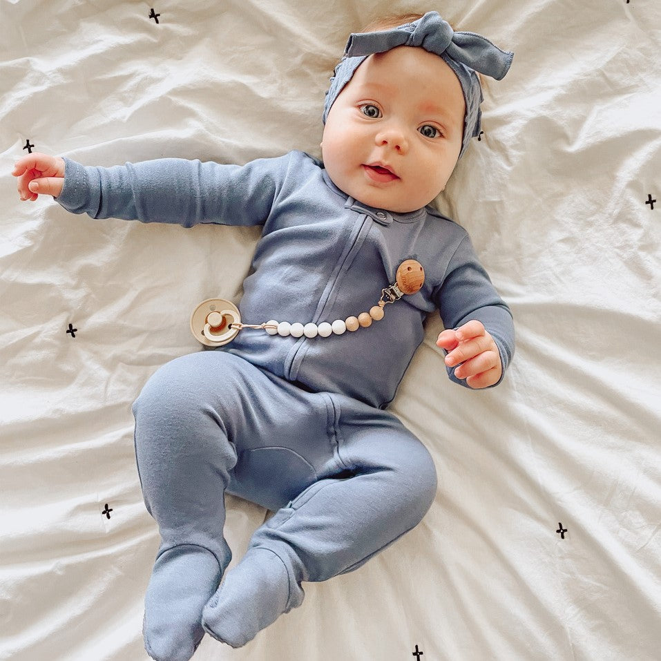 Baby in a blue outfit with a teething necklace lying on a white blanket.