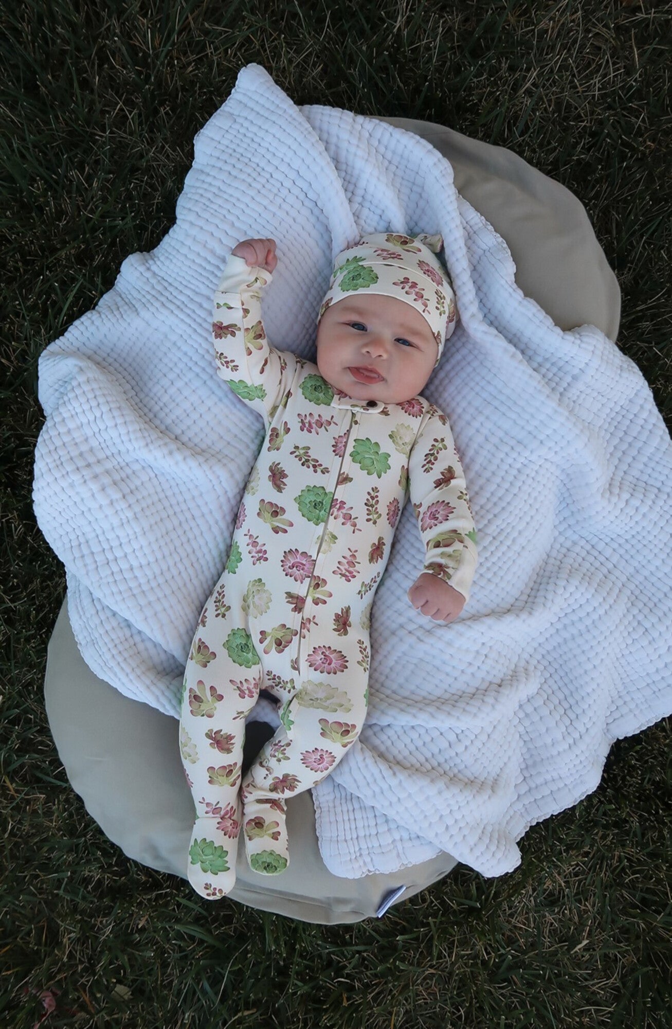 Baby in floral onesie and hat lying on a white blanket outdoors