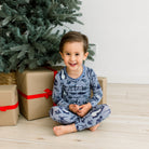A smiling young boy sits cross-legged on a light wood floor in front of a Christmas tree, wearing a blue long-sleeve pajama set with a black space-themed print. Gift boxes are wrapped in brown paper and tied with red ribbons.
