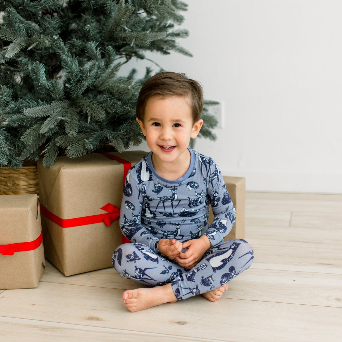 A smiling young boy sits cross-legged on a light wood floor in front of a Christmas tree, wearing a blue long-sleeve pajama set with a black space-themed print. Gift boxes are wrapped in brown paper and tied with red ribbons.