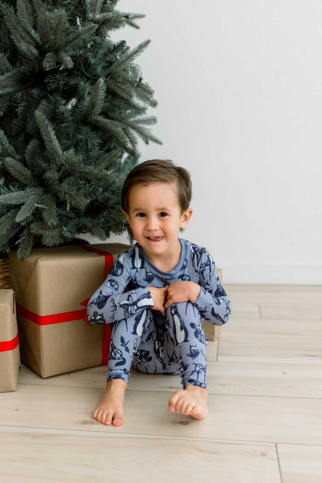 A young child sitting on a light wood floor next to a Christmas tree and wrapped gifts, wearing blue pajamas with a dark blue animal print.