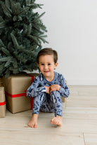 A young child sitting on a light wood floor next to a Christmas tree and wrapped gifts, wearing blue pajamas with a dark blue animal print.