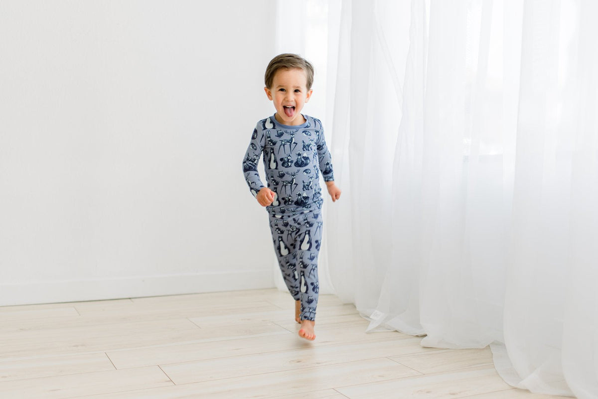 A toddler boy wearing a long-sleeve pajama set with a blue and gray animal print, running barefoot on a light wood floor in front of white curtains.
