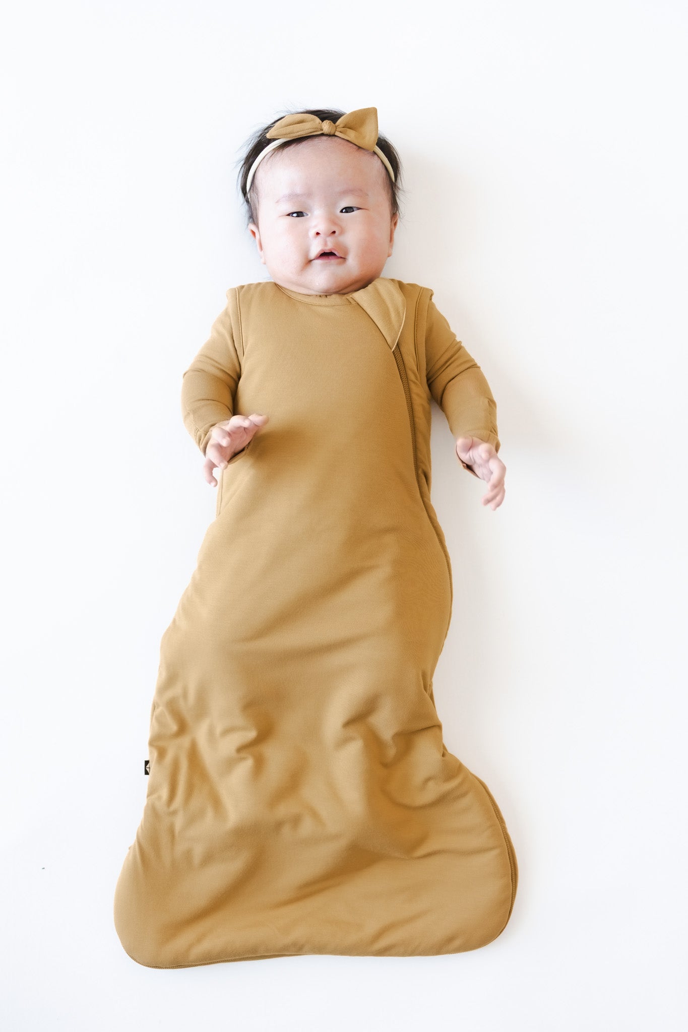 A baby lying on their back wearing a mustard yellow long-sleeve sleep sack and matching bow headband, set against a plain white background.