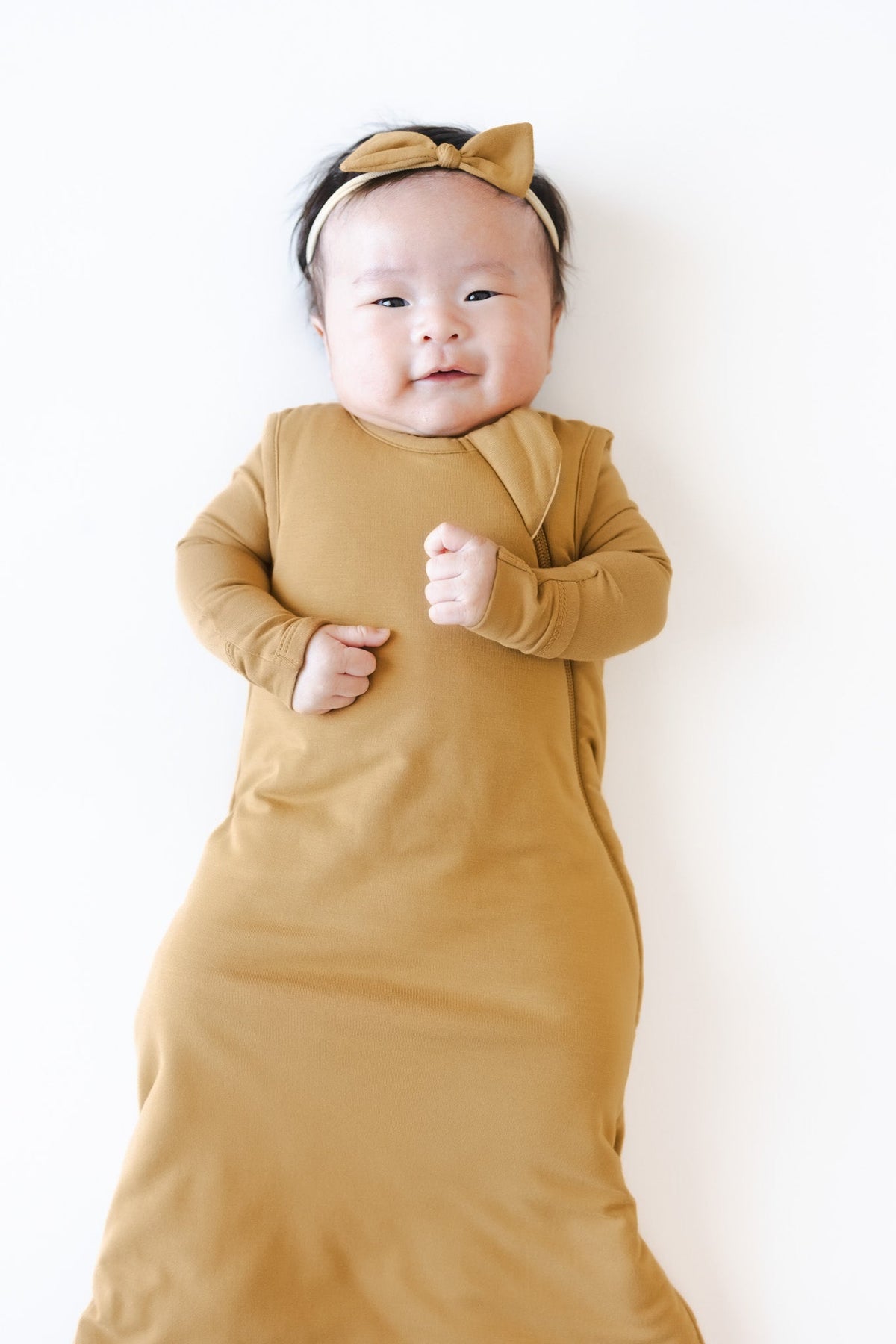 A baby wearing a mustard yellow long-sleeve wrap swaddle and a matching bow headband, lying on a plain white background.