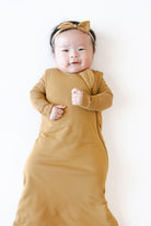 A baby wearing a mustard yellow long-sleeve wrap swaddle and a matching bow headband, lying on a plain white background.