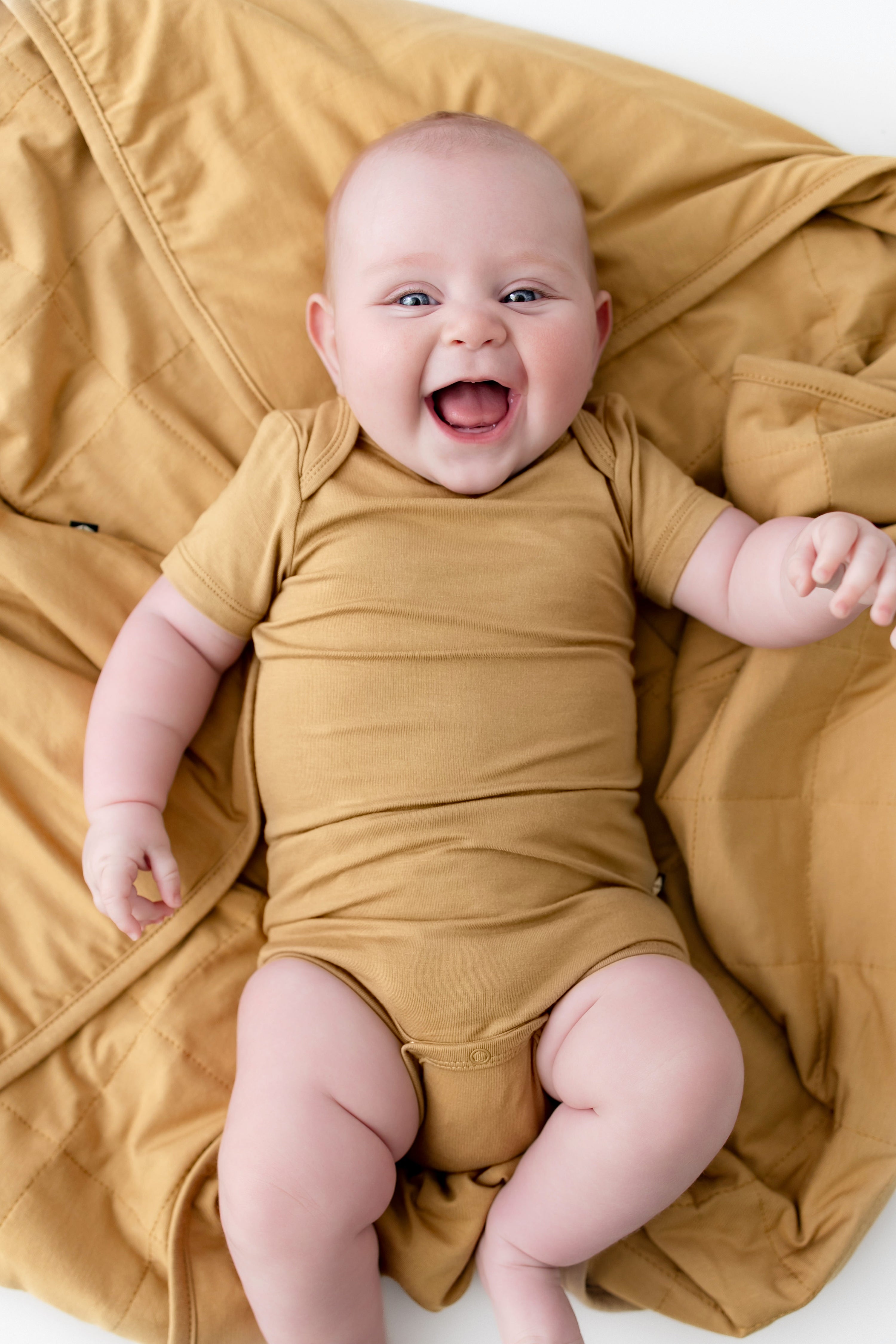 A smiling baby lying on a mustard yellow blanket, wearing a matching mustard yellow short-sleeve bodysuit.