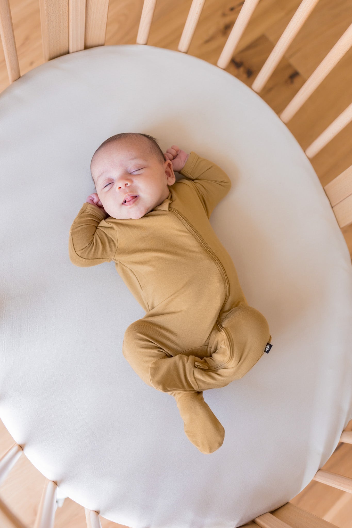 A sleeping newborn baby wearing a mustard yellow footed zip-up sleeper, lying on a white oval mattress inside a wooden crib.