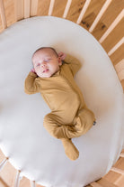 A sleeping newborn baby wearing a mustard yellow footed zip-up sleeper, lying on a white oval mattress inside a wooden crib.