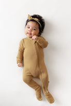 A baby lying on a white background wearing a tan footed zip sleeper and a matching tan headband with a bow.