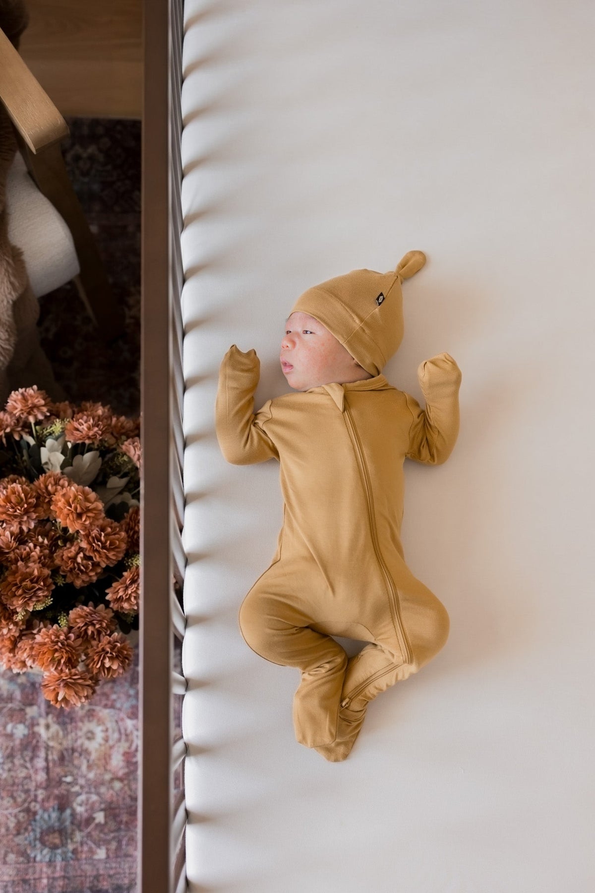 A newborn baby wearing a mustard yellow full-body footed onesie and matching hat, lying on their back on a white textured blanket next to a wooden frame with dried flowers.