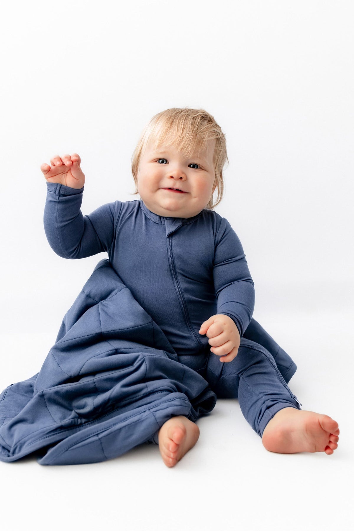 A baby wearing a navy blue long-sleeve footed sleep sack sits on a white studio floor, looking at the camera.